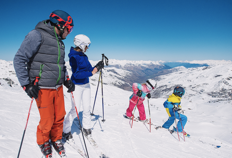 two children are skiing and the parents are watching, infront of a snowy group of mountains. 